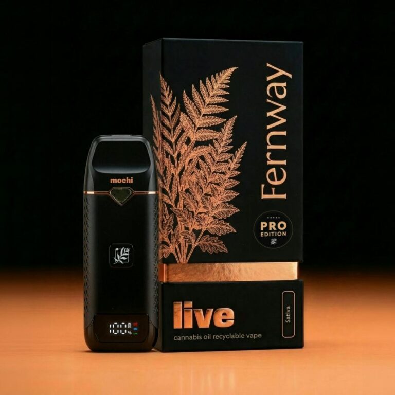 Fernway Live Resin Cartridge – Mochi Product photo of the Fernway Pro Edition Live Cannabis Oil Recyclable Vape in the Mochi strain (Sativa), displayed against a dark background with warm amber lighting. The sleek black vape device features a small digital display showing 100% battery and a rose gold accent ring, with the strain name "mochi" printed near the top. It is positioned in front of its premium black and rose gold packaging, which is embossed with a detailed fern illustration and labeled "Fernway Pro Edition – Live Cannabis Oil Recyclable Vape – Sativa."